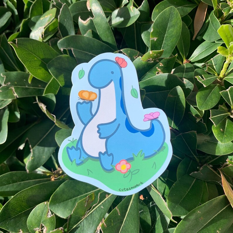 May include: A blue dinosaur sticker with a flower on its head, holding a brown cookie. The dinosaur is sitting on a green patch of grass with pink flowers. The sticker has the text "CutesourArt" at the bottom.