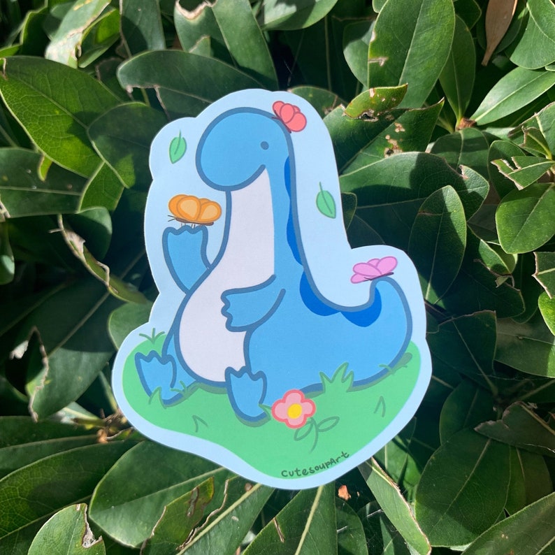 May include: A blue dinosaur sticker with a butterfly and flowers. The sticker has a light blue background and the text "CutesoupArt" at the bottom.