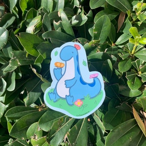 May include: A blue dinosaur sticker with a flower on its head, holding a small orange object. The dinosaur is sitting in a field of green grass with pink flowers. The sticker says "Cutesummer" at the bottom.