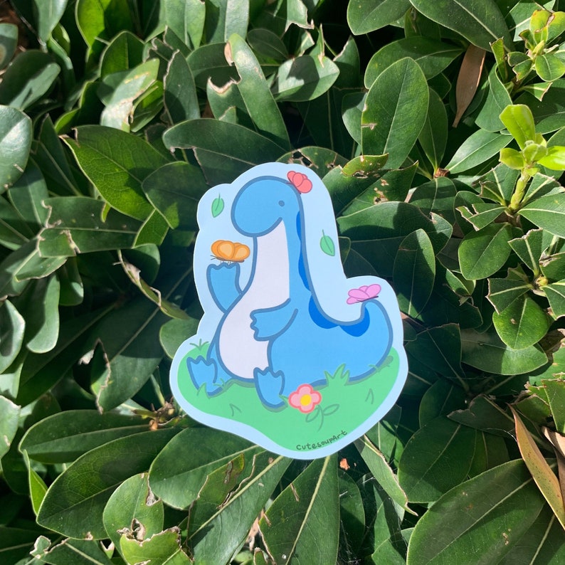 May include: A blue dinosaur sticker with a flower on its head, holding a small orange object. The dinosaur is sitting in a field of green grass with pink flowers. The sticker says "Cutesummer" at the bottom.