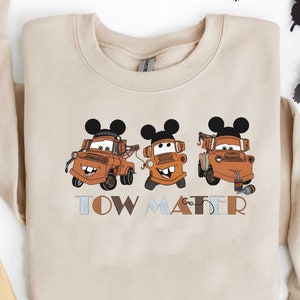 May include: A beige sweatshirt with a graphic of three Tow Mater characters from the Disney Pixar Cars movie. Each character is wearing Mickey Mouse ears. The text "TOW MATER" is printed below the characters.