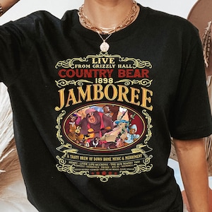 以下が含まれることがあります： グリズリーホールカントリーベアジャンボリー1898の文字と、バンジョーを弾くクマのイラストが描かれた、ヴィンテージスタイルのグラフィックデザインの黒いTシャツ。イラストには、クマの周りに他の動物も描かれている。