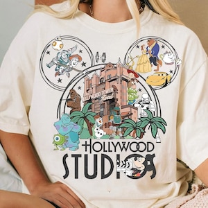 Può includere: Una maglietta bianca con una stampa grafica di una testa di Topolino con vari personaggi e attrazioni Disney al suo interno. Il testo "HOLLYWOOD STUDIOS" è stampato sotto la grafica.