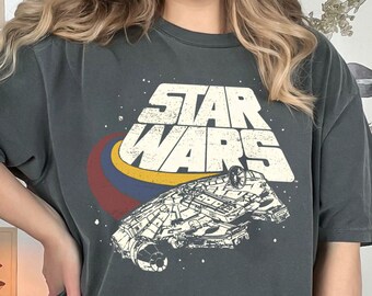 以下が含まれることがあります： 暗いグレーのTシャツに、ミレニアム・ファルコンの宇宙船と「STAR WARS」の文字がヴィンテージスタイルで描かれています。文字は白と黄色です。