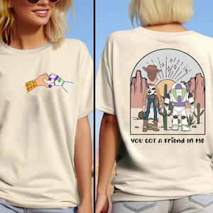May include: Two white t-shirts with a cartoon image of Woody and Buzz Lightyear from Toy Story. The front shirt shows Woody and Buzz holding hands in a heart shape. The back shirt shows Woody and Buzz standing together in a desert landscape with the text "You got a friend in me".