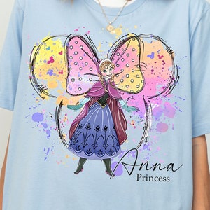 Peut inclure: T-shirt bleu clair avec un motif color&eacute; d'Anna de La Reine des Neiges portant un n&oelig;ud rose et jaune. Le motif est entour&eacute; d'&eacute;claboussures de peinture color&eacute;es. Le texte "Anna Princess" est imprim&eacute; sous le motif.