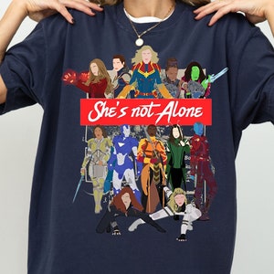 Puede incluir: Una camiseta azul marino con un gráfico de un grupo de superheroínas. El gráfico incluye el texto "She's not Alone".