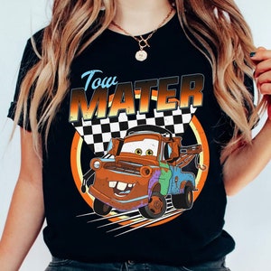 May include: Black t-shirt featuring a cartoon image of Tow Mater from the Cars movie. The design includes the text "Tow Mater" in a stylized font, a checkered flag, and a circular orange and yellow background. The graphic is centered on the shirt.