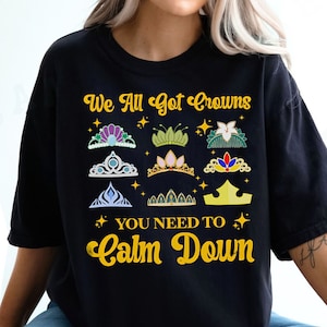 Op de afbeelding: Zwart T-shirt met een gouden afbeelding met de tekst "We All Got Crowns You Need To Calm Down". De afbeelding bevat een verscheidenheid aan kleurrijke kronen.
