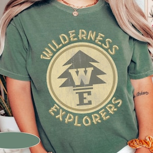 Puede incluir: Una camiseta verde con un gráfico de estilo vintage de un pino y el texto "Wilderness Explorers" en un círculo.