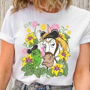 May include: White t-shirt featuring a cartoon illustration of a horse and a chameleon, surrounded by flowers. The horse wears a black hat with the word "Maximus" on it. The chameleon wears a black hat with the word "Pascal" on it.