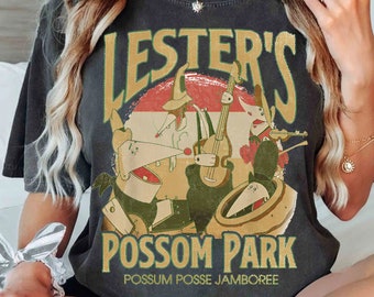 May include: Dark gray t-shirt featuring the text "LESTER'S POSSOM PARK" in gold lettering with a red and white circular background. The graphic depicts cartoon possums playing musical instruments. Below the text is "POSSUM POSSE JAMBOREE".