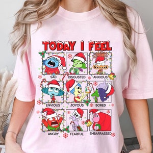 May include: Light pink t-shirt featuring the phrase "TODAY I FEEL" above nine cartoon characters, each representing a different emotion. The characters are wearing Santa hats and surrounded by Christmas lights and candy canes. Emotions include sad, disgusted, anxious, envious, joyous, bored, angry, fearful, and embarrassed.