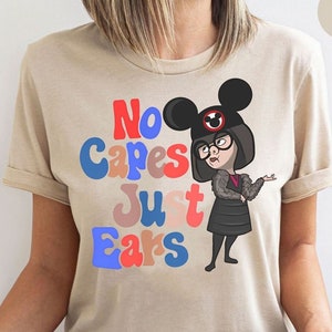 May include: A beige t-shirt with a colorful graphic of a woman wearing a black dress and Mickey Mouse ears. The text "No capes just ears" is printed on the shirt in a retro font.