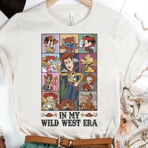 Puede incluir: Una camiseta blanca con un gr&aacute;fico de dibujos animados de varios personajes de Disney, incluidos Woody, Buzz Lightyear y Mickey Mouse. El gr&aacute;fico est&aacute; rodeado por un borde negro y el texto "In My Wild West Era" est&aacute; impreso debajo del gr&aacute;fico.