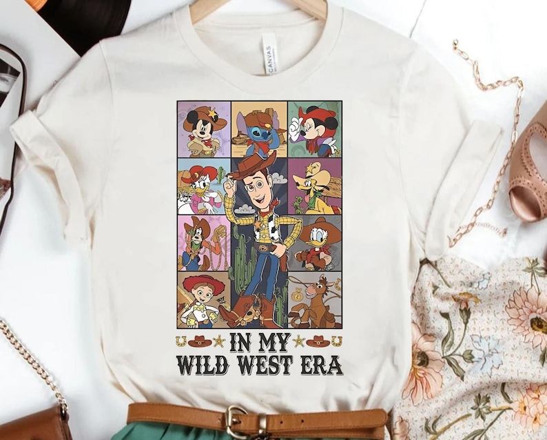 Puede incluir: Una camiseta blanca con un gr&aacute;fico de dibujos animados de varios personajes de Disney, incluidos Woody, Buzz Lightyear y Mickey Mouse. El gr&aacute;fico est&aacute; rodeado por un borde negro y el texto "In My Wild West Era" est&aacute; impreso debajo del gr&aacute;fico.