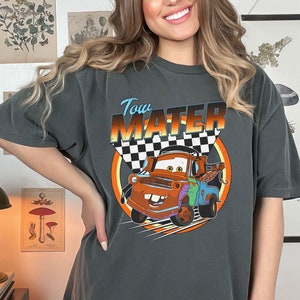 May include: Dark gray t-shirt featuring the character Tow Mater from the movie Cars. The design includes the text "Tow Mater" above an image of the character with a checkered flag background. The shirt is a relaxed fit.