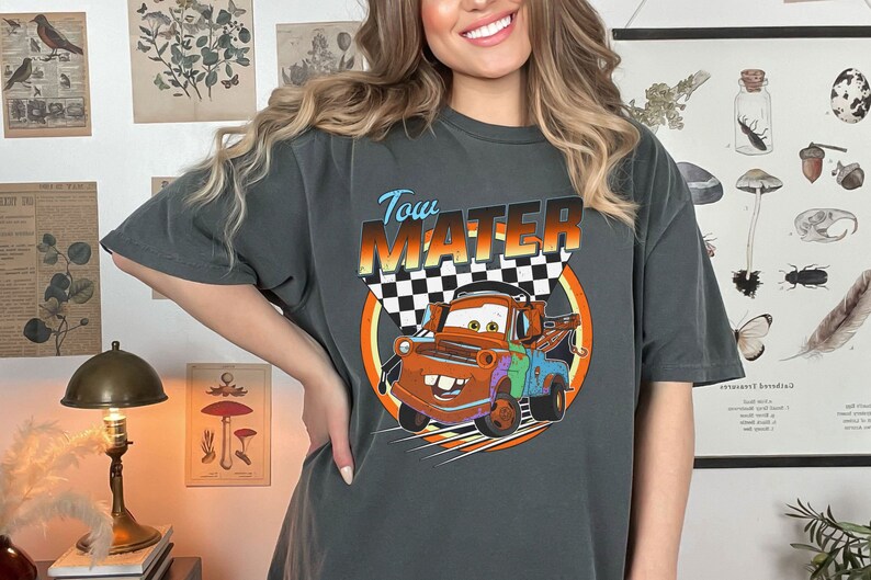 May include: Dark gray t-shirt featuring the character Tow Mater from the movie Cars. The design includes the text "Tow Mater" above an image of the character with a checkered flag background. The shirt is a relaxed fit.