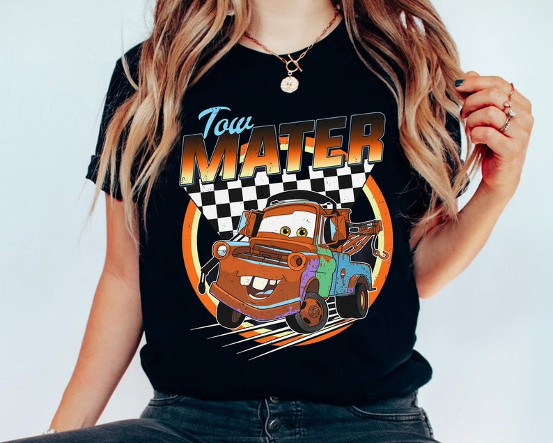 May include: Black t-shirt featuring a cartoon image of Tow Mater from the Cars movie. The design includes the text "Tow Mater" in a stylized font, a checkered flag, and a circular orange and yellow background. The graphic is centered on the shirt.