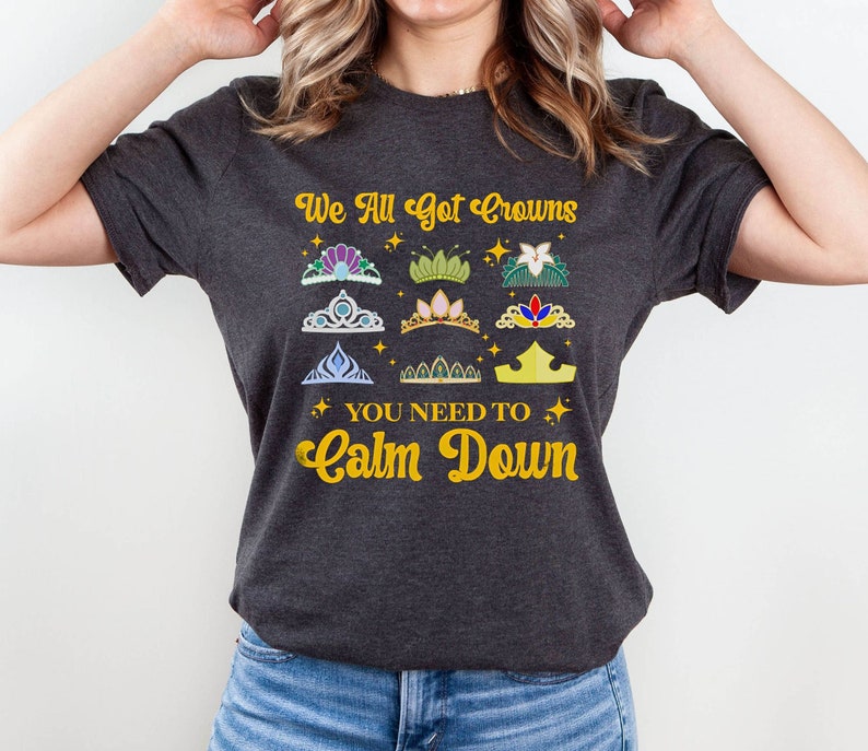 Op de afbeelding: Een donkergrijs T-shirt met de tekst "We All Got Crowns You Need To Calm Down" in gouden letters. Het shirt heeft een verscheidenheid aan kleurrijke kronen met gouden accenten.