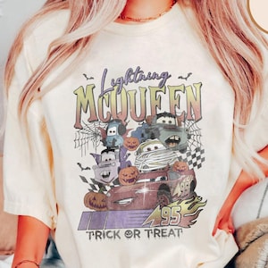 May include: Cream-colored t-shirt featuring a Halloween-themed graphic of Lightning McQueen and other Cars characters. The design includes pumpkins, bats, spiderwebs, and the text "Lightning McQueen Trick or Treat".