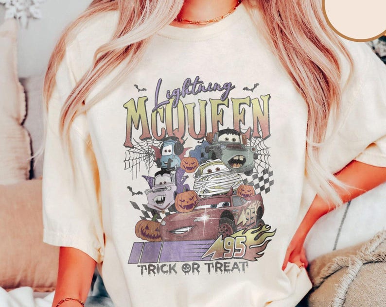 May include: Cream-colored t-shirt featuring a Halloween-themed graphic of Lightning McQueen and other Cars characters. The design includes pumpkins, bats, spiderwebs, and the text "Lightning McQueen Trick or Treat".