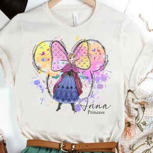 Peut inclure: Un t-shirt blanc avec un motif aquarelle d'Anna de La Reine des Neiges. Le motif repr&eacute;sente Anna dans une robe violette avec un n&oelig;ud rose et le texte "Anna Princess".