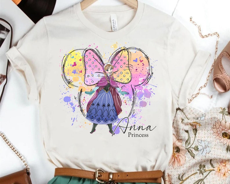 Peut inclure: Un t-shirt blanc avec un motif aquarelle d'Anna de La Reine des Neiges. Le motif repr&eacute;sente Anna dans une robe violette avec un n&oelig;ud rose et le texte "Anna Princess".