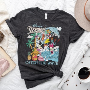 May include: A dark gray t-shirt with a colorful graphic of Mickey Mouse and friends at Disney's Typhoon Lagoon water park. The graphic includes the text "Disney's Typhoon Lagoon" and "Catch the Wave".