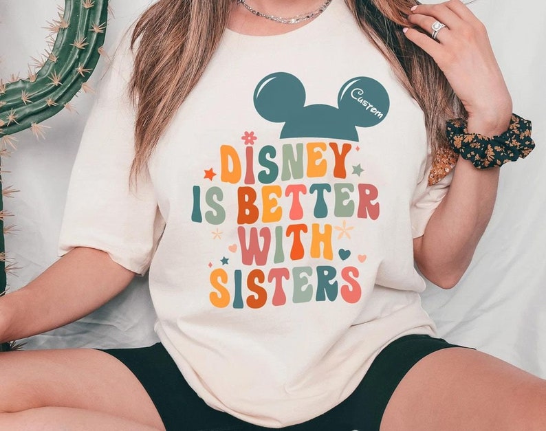 May include: A white t-shirt with a rainbow-colored graphic that says "Disney is better with sisters" and a black Mickey Mouse head with the word "Custom" above it.