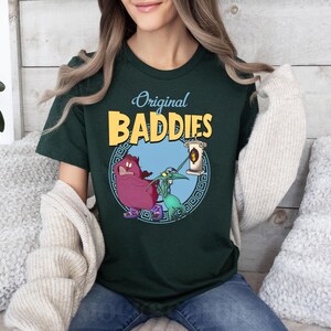 May include: A dark green t-shirt featuring a graphic print of two cartoon characters. The characters are a purple creature with a crown and a green creature with a long nose. The text "Original Baddies" is printed in yellow above the characters.
