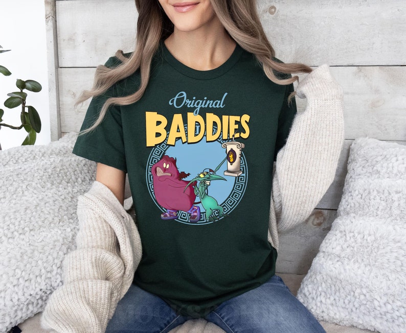 May include: A dark green t-shirt featuring a graphic print of two cartoon characters. The characters are a purple creature with a crown and a green creature with a long nose. The text "Original Baddies" is printed in yellow above the characters.