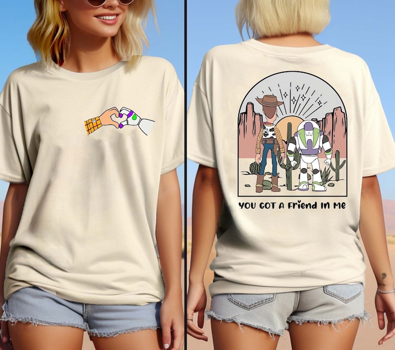 May include: Two white t-shirts with a cartoon image of Woody and Buzz Lightyear from Toy Story. The front shirt shows Woody and Buzz holding hands in a heart shape. The back shirt shows Woody and Buzz standing together in a desert landscape with the text "You got a friend in me".