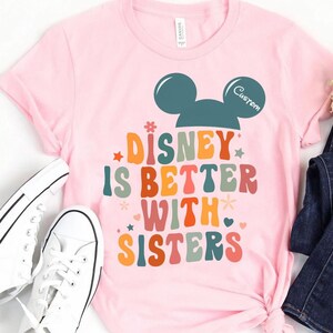 May include: Pink t-shirt with a retro-style graphic that says "Disney is better with sisters" and a Mickey Mouse head silhouette. The Mickey Mouse head is teal blue and the text is in rainbow colors.