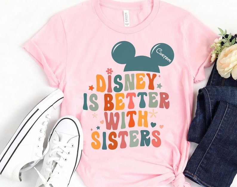 May include: Pink t-shirt with a retro-style graphic that says "Disney is better with sisters" and a Mickey Mouse head silhouette. The Mickey Mouse head is teal blue and the text is in rainbow colors.
