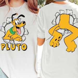 Könnte beinhalten: Weißes T-Shirt mit einem Cartoon-Bild von Pluto dem Hund, der aus einem zerrissenen Loch schaut. Der Text "PLUTO" ist unter dem Bild gedruckt. Die Rückseite des Shirts zeigt Plutos Beine, die aus einem zerrissenen Loch ragen.