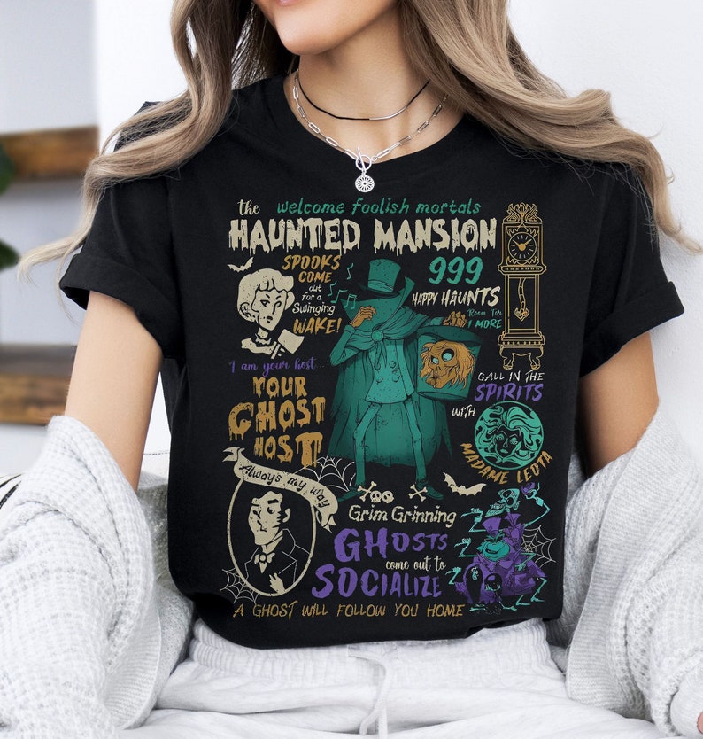 May include: Black t-shirt with a colorful graphic design featuring a haunted mansion theme. The design includes text that says "The Welcome Foolish Mortals Haunted Mansion 999 Happy Haunts", "Your Ghost Host", "Grim Grinning Ghosts Come Out To Socialize", and "A Ghost Will Follow You Home".