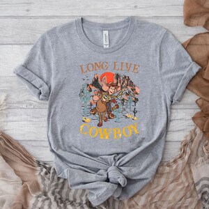 Pu&ograve; includere: Maglietta grigia con una grafica di Woody e Bullseye di Toy Story che cavalcano attraverso un paesaggio di tramonto. Il testo "Long Live Cowboy" &egrave; stampato sulla maglietta.