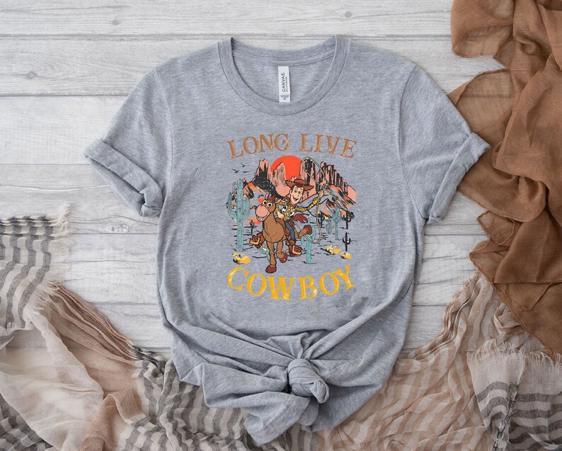 Pu&ograve; includere: Maglietta grigia con una grafica di Woody e Bullseye di Toy Story che cavalcano attraverso un paesaggio di tramonto. Il testo "Long Live Cowboy" &egrave; stampato sulla maglietta.