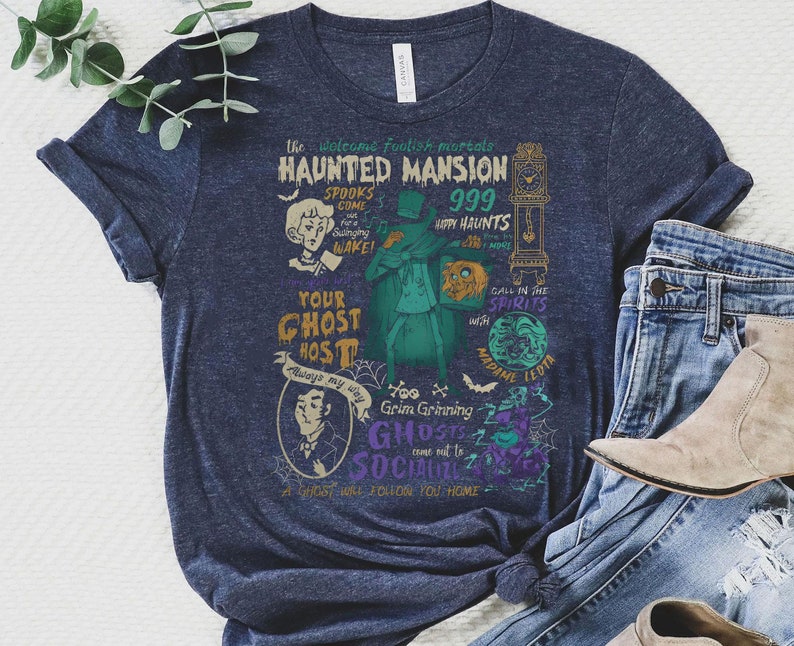 May include: A navy blue t-shirt with a vintage-style graphic design featuring a haunted mansion, ghosts, and the text "The Welcome Foolish Mortals Haunted Mansion 999 Happy Haunts".