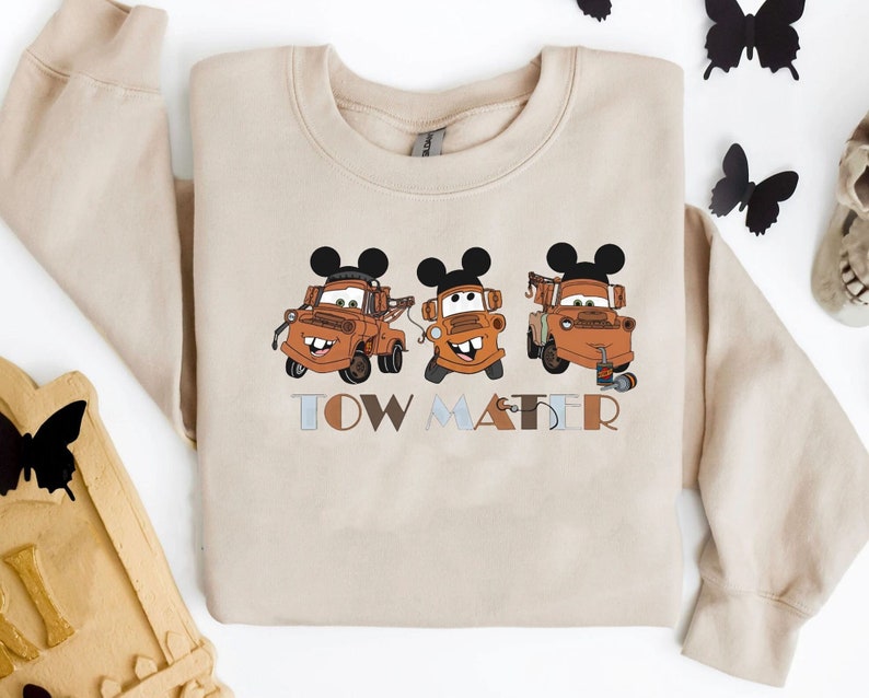 May include: A beige sweatshirt with a graphic of three Tow Mater characters from the Disney Pixar Cars movie. Each character is wearing Mickey Mouse ears. The text "TOW MATER" is printed below the characters.