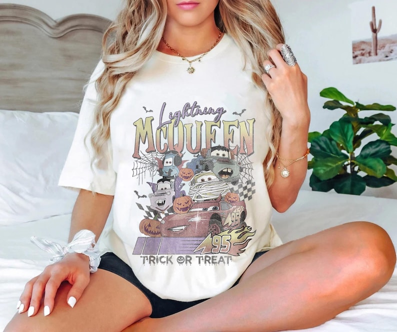 May include: Cream-colored t-shirt featuring a Halloween-themed graphic of Lightning McQueen and other Cars characters. The design includes pumpkins, bats, and the text "Lightning McQueen Trick or Treat" in a vintage style.