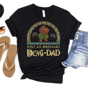 May include: Black t-shirt featuring a cartoon character and the text "JUST AN ORDINARY DEMI-DAD". The image includes denim shorts, sunglasses, and sandals. A black circle is in the upper left corner.