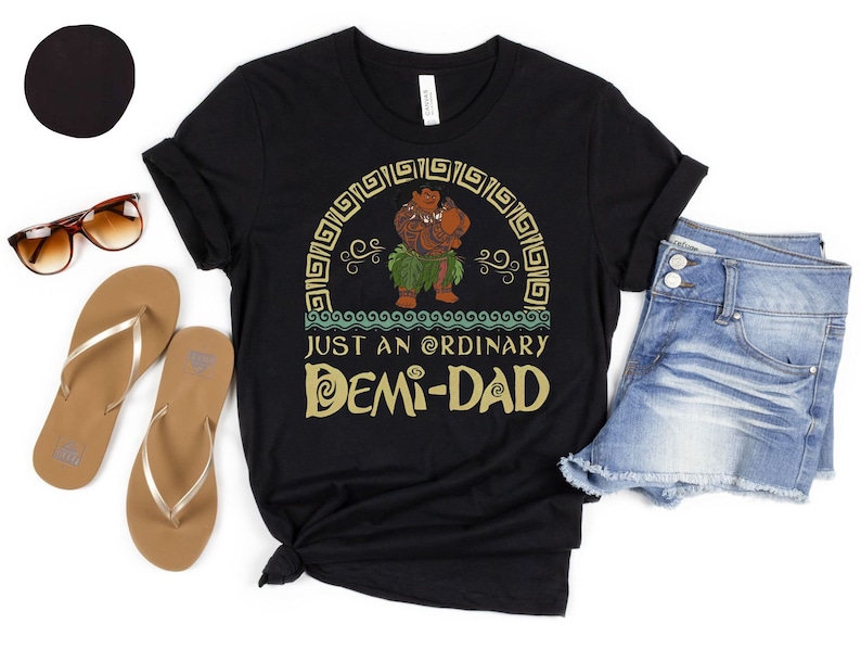 May include: Black t-shirt featuring a cartoon character and the text "JUST AN ORDINARY DEMI-DAD". The image includes denim shorts, sunglasses, and sandals. A black circle is in the upper left corner.
