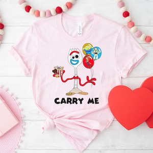 May include: Pink t-shirt with a cartoon image of Forky from Toy Story holding a box of french fries and balloons. The text "Carry Me" is printed below the image.