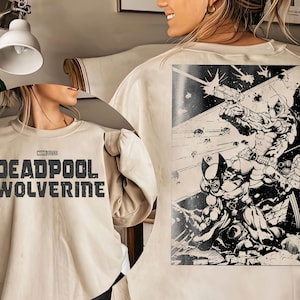 May include: A beige sweatshirt with a black graphic print of Deadpool and Wolverine fighting on the back. The front of the sweatshirt has the text "MARVEL LEGENDS DEADPOOL & WOLVERINE" in black.
