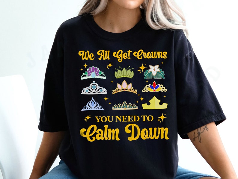 Op de afbeelding: Zwart T-shirt met een gouden afbeelding met de tekst "We All Got Crowns You Need To Calm Down". De afbeelding bevat een verscheidenheid aan kleurrijke kronen.