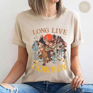 Pu&ograve; includere: Una t-shirt beige con una stampa grafica di Woody di Toy Story a cavallo in un paesaggio desertico. Il testo "Long Live Cowboy" &egrave; stampato sulla t-shirt.