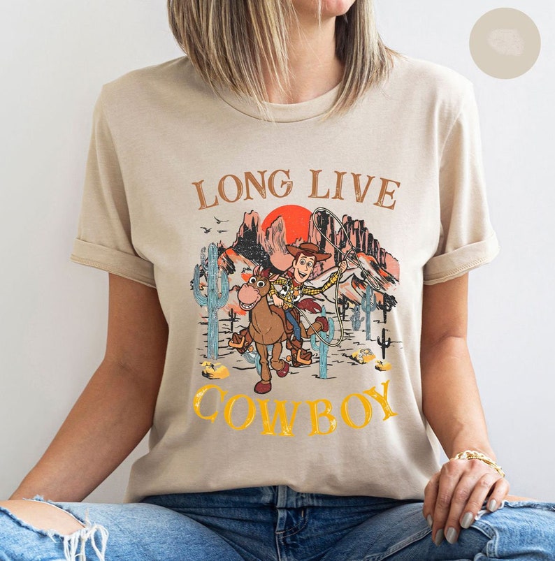 Pu&ograve; includere: Una t-shirt beige con una stampa grafica di Woody di Toy Story a cavallo in un paesaggio desertico. Il testo "Long Live Cowboy" &egrave; stampato sulla t-shirt.