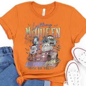 May include: Orange t-shirt featuring a graphic of Lightning McQueen and other Cars characters in Halloween costumes. The text "Lightning McQueen" and "Trick or Treat" are also displayed. The shirt is paired with blue jeans and white sneakers.